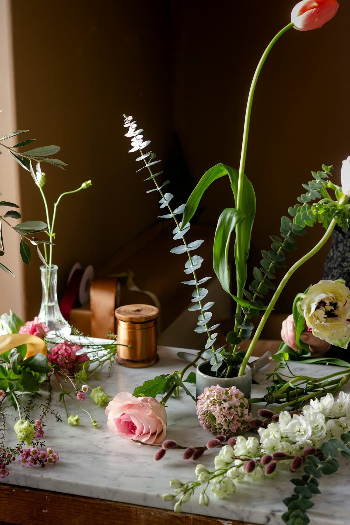 Florist workbench mid-arrangement: tulips, eucalyptus, garden roses, and ribbon spools on marble